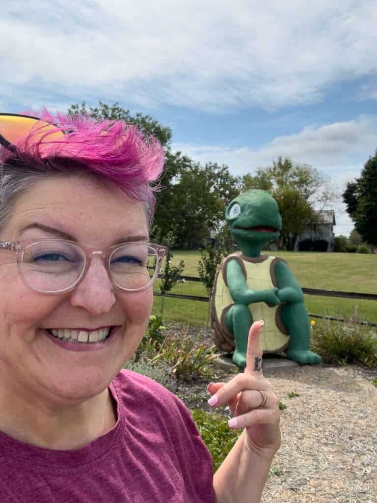 Colorful grandma with pink hair and glasses smiling at camera in natural outdoor setting.Monster Turtle statue at Fulk Lake in Churubusco, Indiana, inspired by local legend
