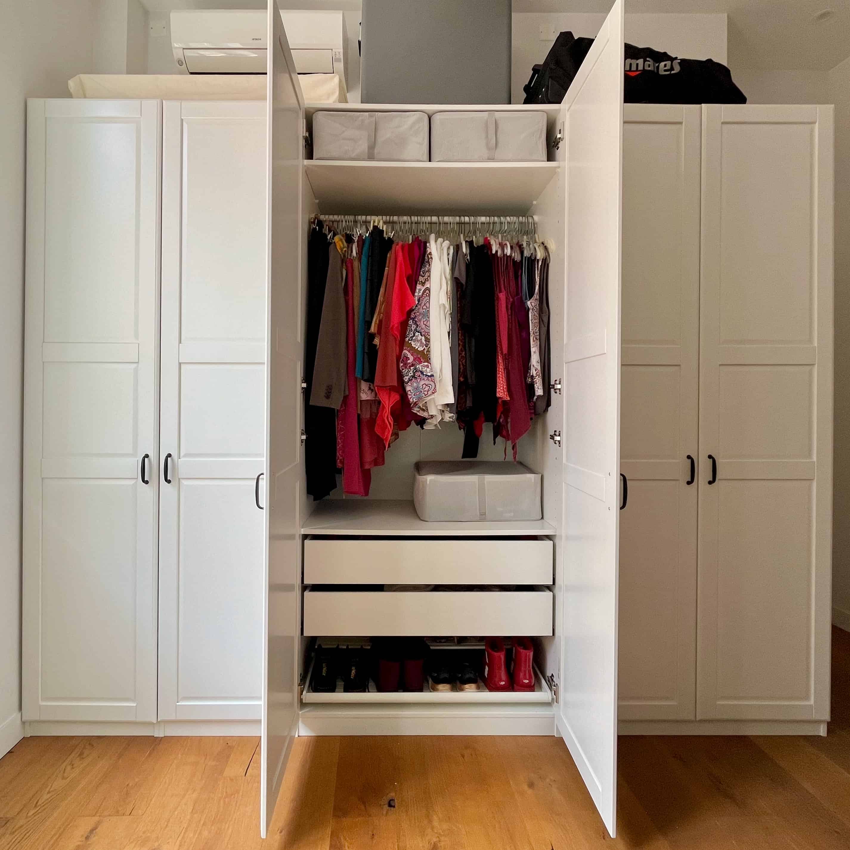 Photo of a wide closet with tall white doors with the middle doors open to show an organized interior of hanging clothes, bins, drawers, and shoes