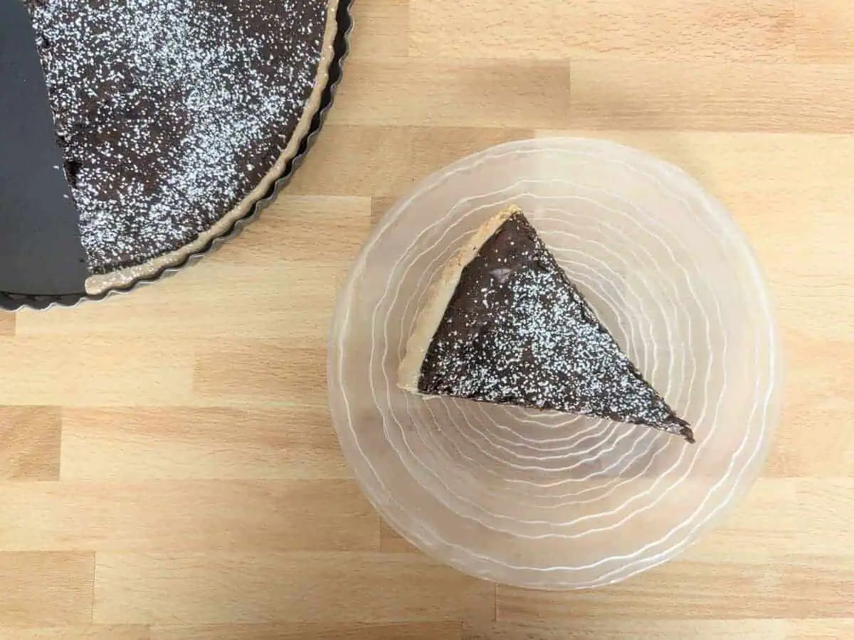 A slice of chocolate tart on a clear plate with a fork. The tart is topped with powdered sugar.