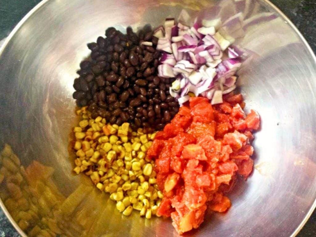 A metal bowl containing separate piles of black beans, chopped red onion, corn, and diced tomatoes—the fresh ingredients ready to be mixed into a vibrant black bean and corn salsa.