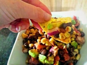 A hand holds a tortilla chip topped with vibrant black bean and corn salsa—featuring avocado, red onion, and tomato—over a bowl brimming with the same flavorful mixture.