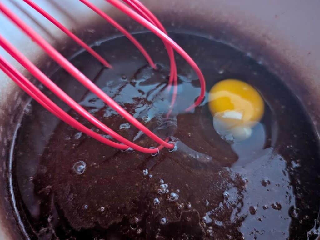 A red whisk mixing a raw egg into a bowl of dark, liquid batter for delicate lacy cookies.