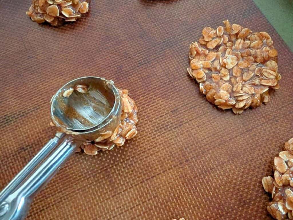 A metal scoop sits on a textured brown surface next to clusters of uncooked oat and nut mixture, ready to be shaped into delicate lacy cookies.