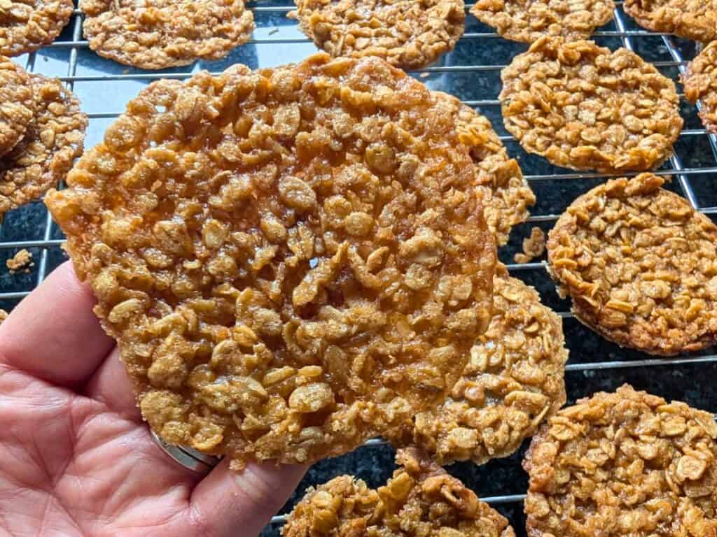 A hand holds a thin, round lacy cookie above a cooling rack, with more delicate lacy cookies in the background.