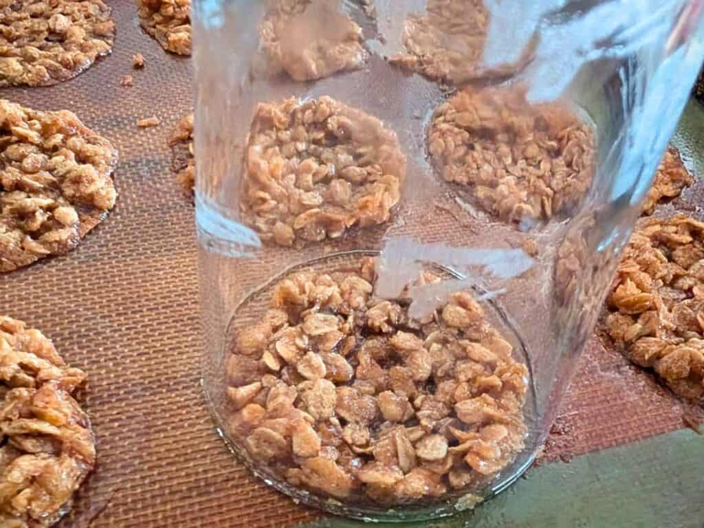 A glass jar is being used to flatten oat lacy cookies on a baking mat, with several unflattened cookies nearby.