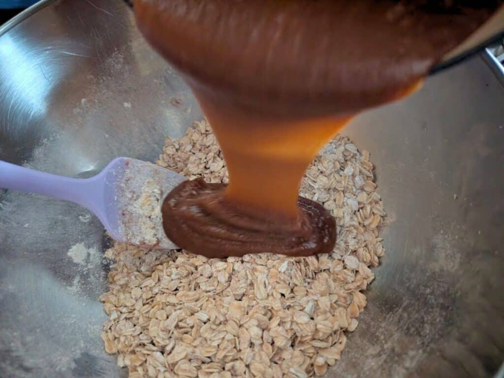 Thick mixture is being poured onto rolled oats in a metal mixing bowl with a spatula, the perfect start for making delicious lacy cookies.