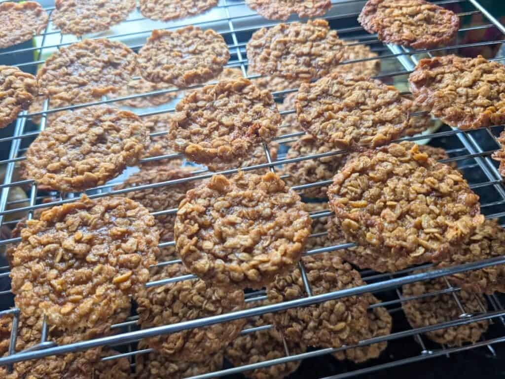Lacy cookies and oatmeal cookies cooling on stacked wire racks, with several rows of golden-brown treats visible in close-up.