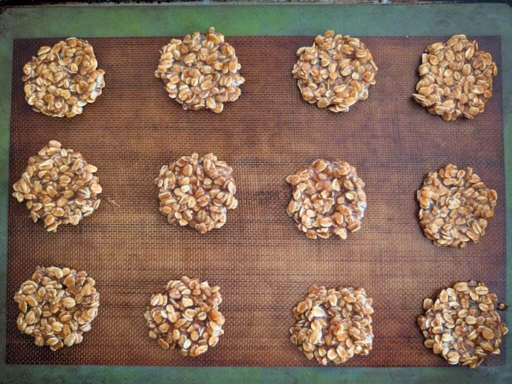 Twelve unbaked lacy cookies, resembling oatmeal cookies, are arranged in rows on a brown baking mat placed on a metal baking sheet.