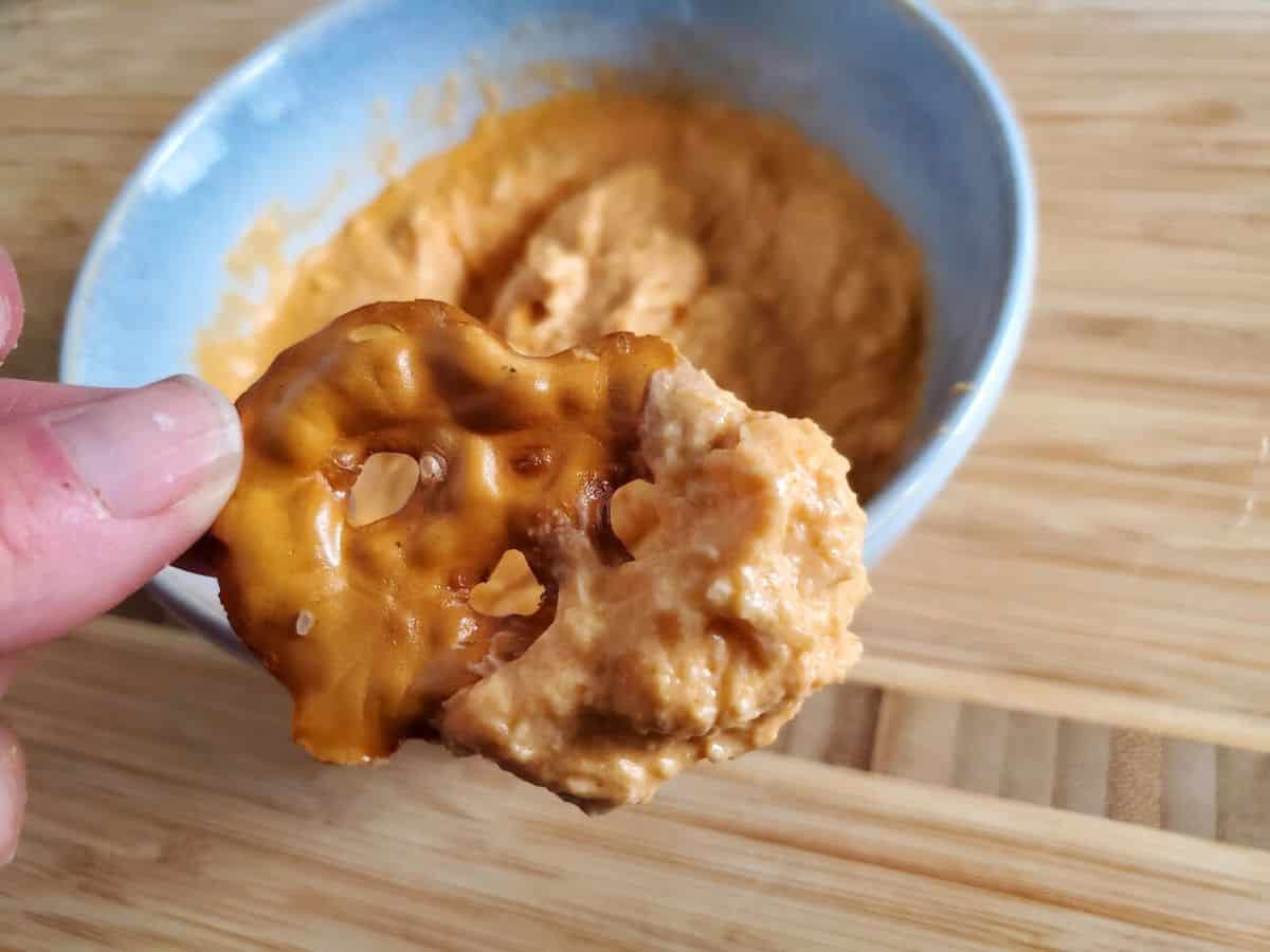Close-up of a hand holding a pretzel chip dipped in a creamy, orange-colored cheese spread. A bowl of the buffalo chicken dip is in the background.