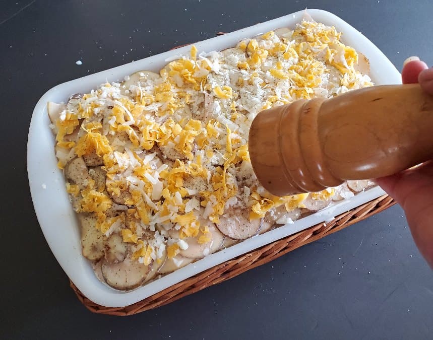 Grating pepper onto unbaked tray of cheesy scalloped potatoes