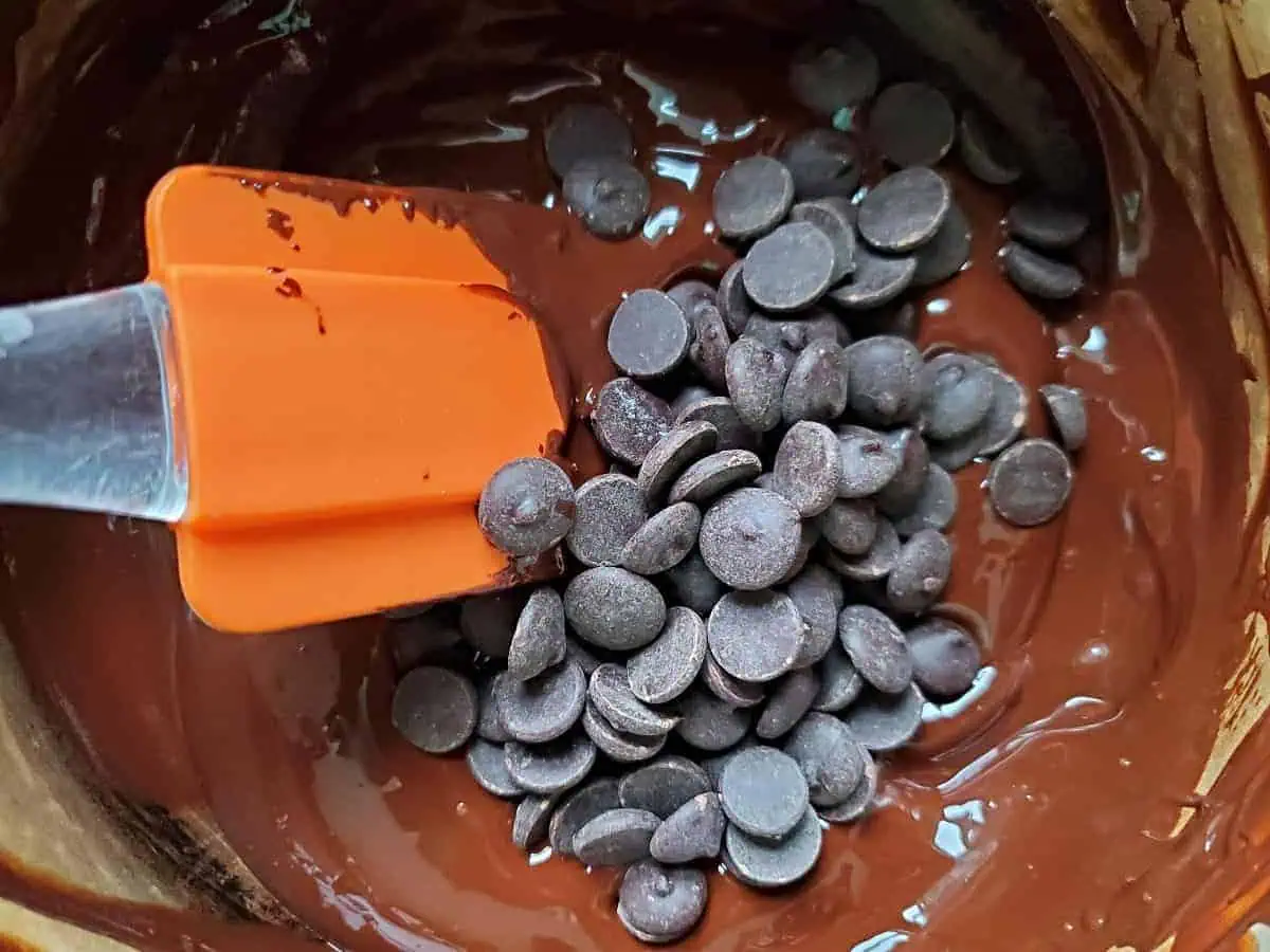 Chocolate callets added to melted chocolate with an orange spatula.