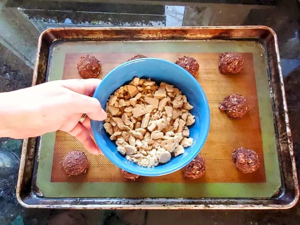 A hand holds a blue bowl of chopped nuts above a baking sheet lined with nine round chocolate dough balls on a silicone mat, ready to become chewy s&rsquo;mores cookies.