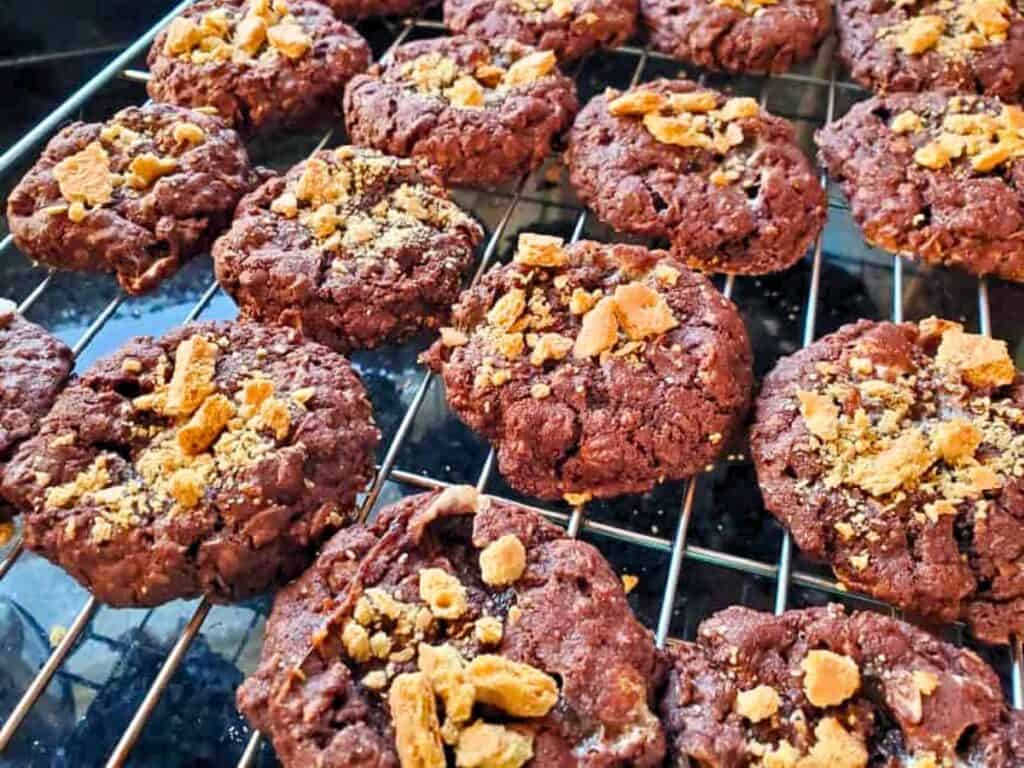 Chewy s&rsquo;mores cookies topped with crushed graham crackers are cooling on a metal wire rack.