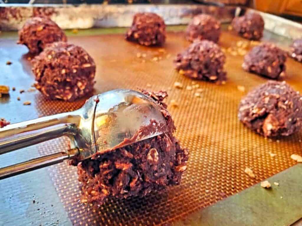 A cookie scoop places chewy s&rsquo;mores cookies dough onto a baking sheet lined with a silicone mat, with several dough balls arranged in the background.
