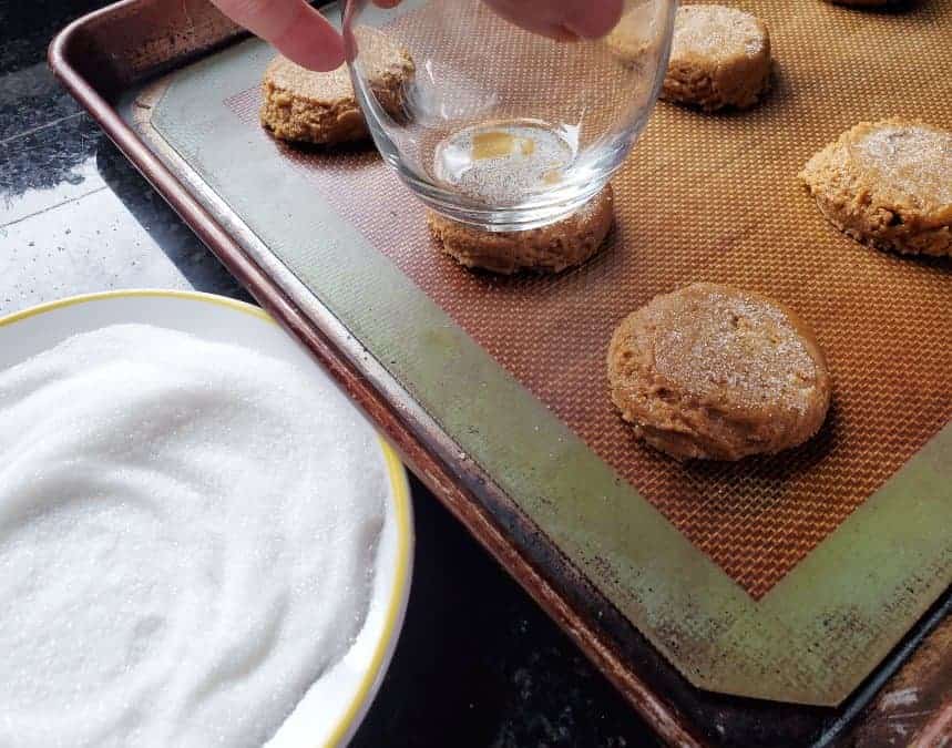 A hand presses a glass onto chewy molasses cookie dough balls on a baking sheet lined with a silicone mat, with a bowl of sugar nearby.