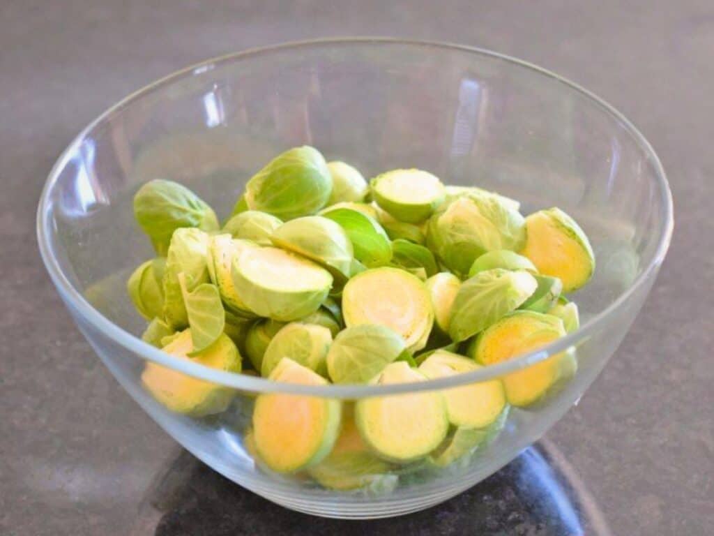 A clear glass bowl filled with halved and whole Brussels sprouts, perfect for making bacon roasted Brussels sprouts, sits on a dark countertop.