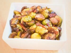 A white square bowl filled with bacon roasted Brussels sprouts sits on a light wooden surface.