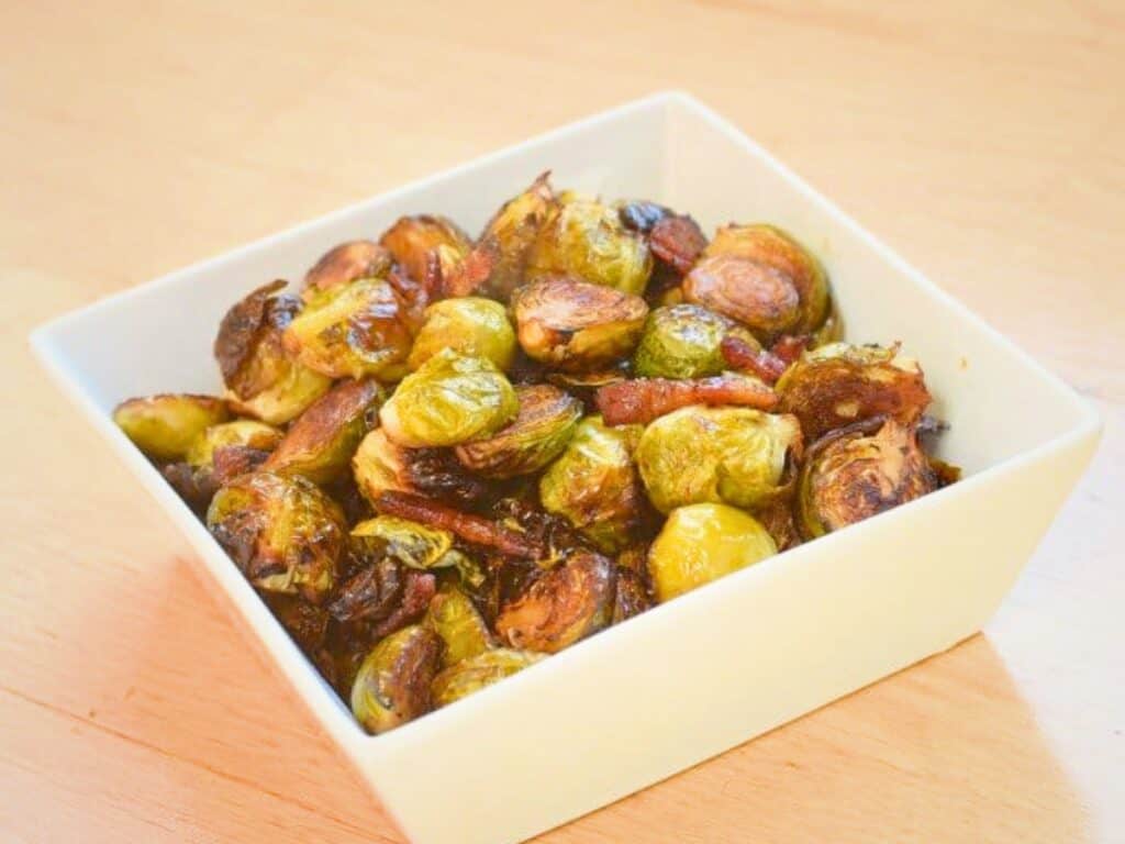 A square white bowl filled with bacon roasted Brussels sprouts and crispy bacon pieces sits on a wooden surface.