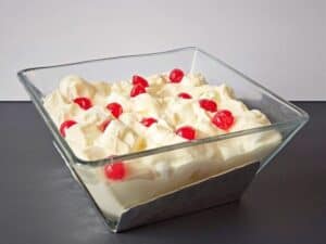 Glass bowl filled with creamy marshmallow salad, topped with several red maraschino cherries, set against a plain background.