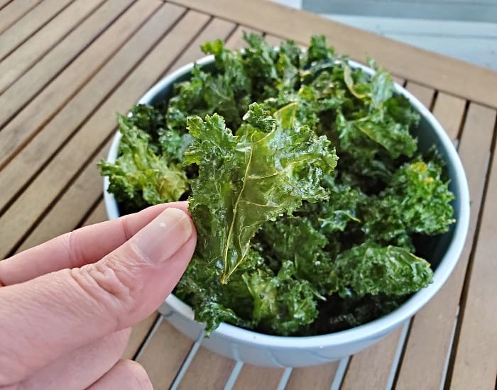 Image shows a hand holding a baked kale chip over a bowl filled with more kale chips.