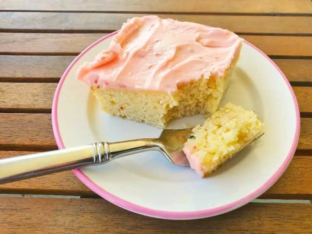 A square piece of yellow cake with pink frosting sits on a white plate, with a fork holding a bite-sized piece.