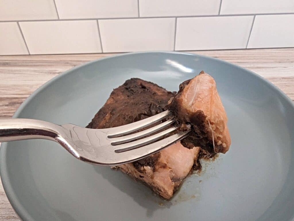 Fork holding a bite of asian marinated salmon with the rest of the fillet on a blue plate in the background.