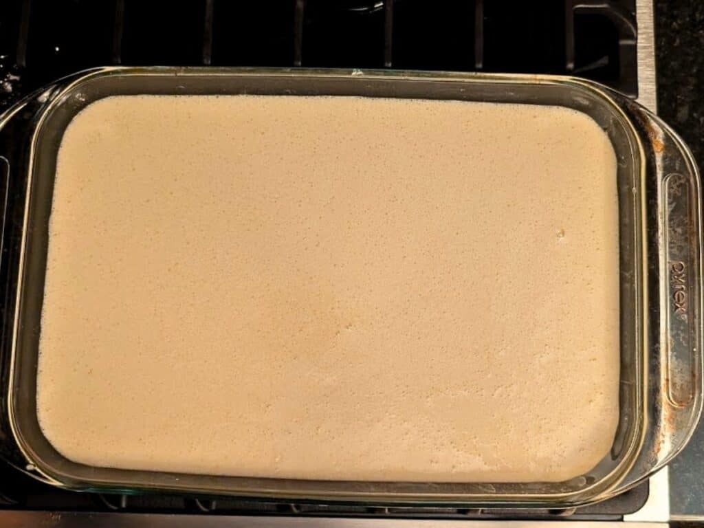 A rectangular glass baking dish filled with a pale, smooth baked lemonade batter sits on a stovetop.
