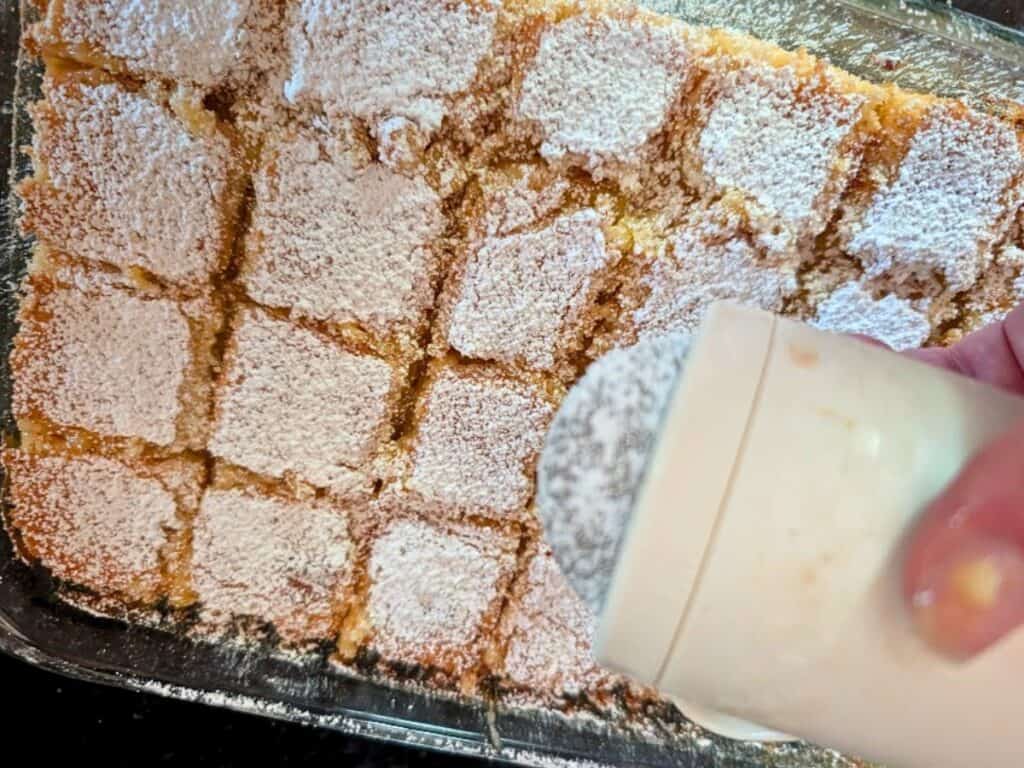 A hand sprinkles powdered sugar over a glass dish of sliced baked lemonade bars.