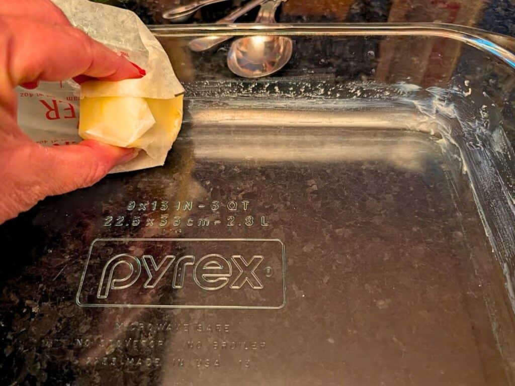 A hand is rubbing a stick of butter on a clear Pyrex baking dish to grease it for baked lemonade, with measuring spoons visible in the background.