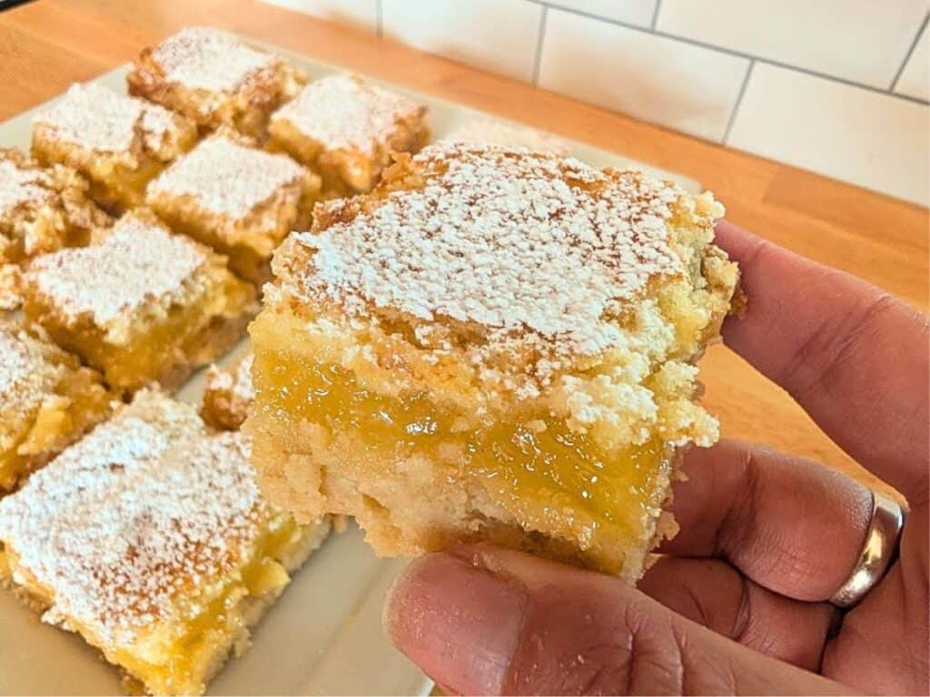 A hand holds a square lemon bar dusted with powdered sugar; more baked lemonade bars are arranged on a plate in the background.