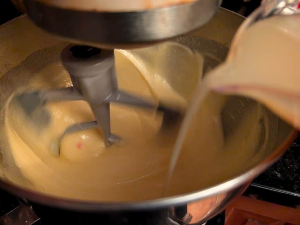 A stand mixer blending batter for baked lemonade while liquid is being poured into the mixing bowl from a measuring cup.