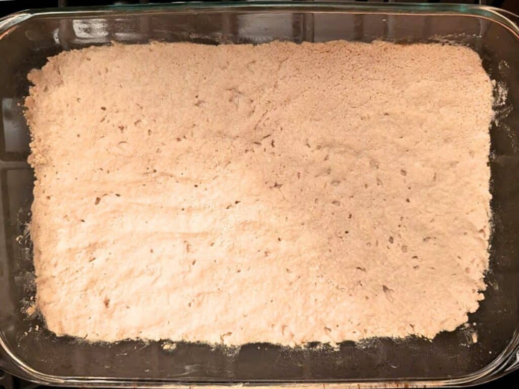 A glass baking dish filled with an even layer of raw, light brown dough, ready to be baked into a sweet treat with a hint of baked lemonade flavor.