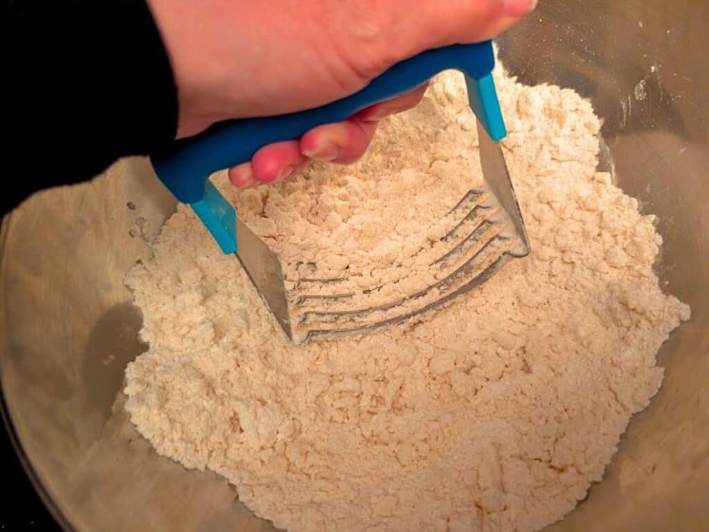 A hand uses a blue-handled pastry cutter to mix flour and fat in a stainless steel bowl, preparing the base for delicious baked lemonade treats.