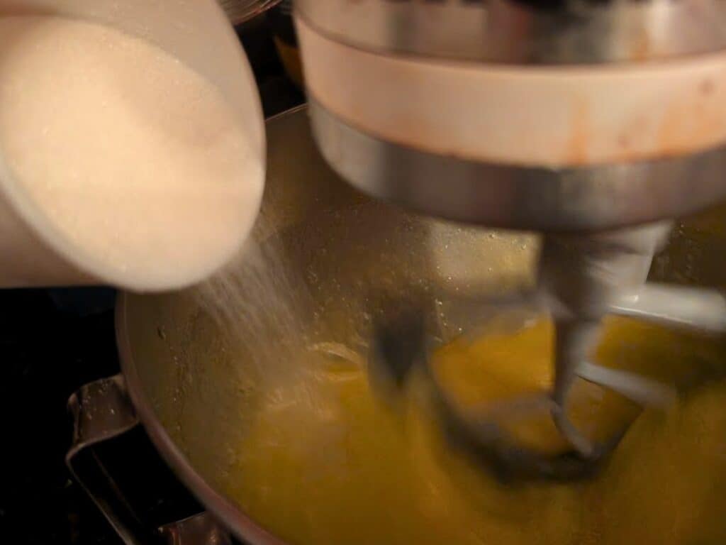 Granulated sugar is being poured into a stand mixer bowl containing a yellow baked lemonade mixture while the mixer is running.