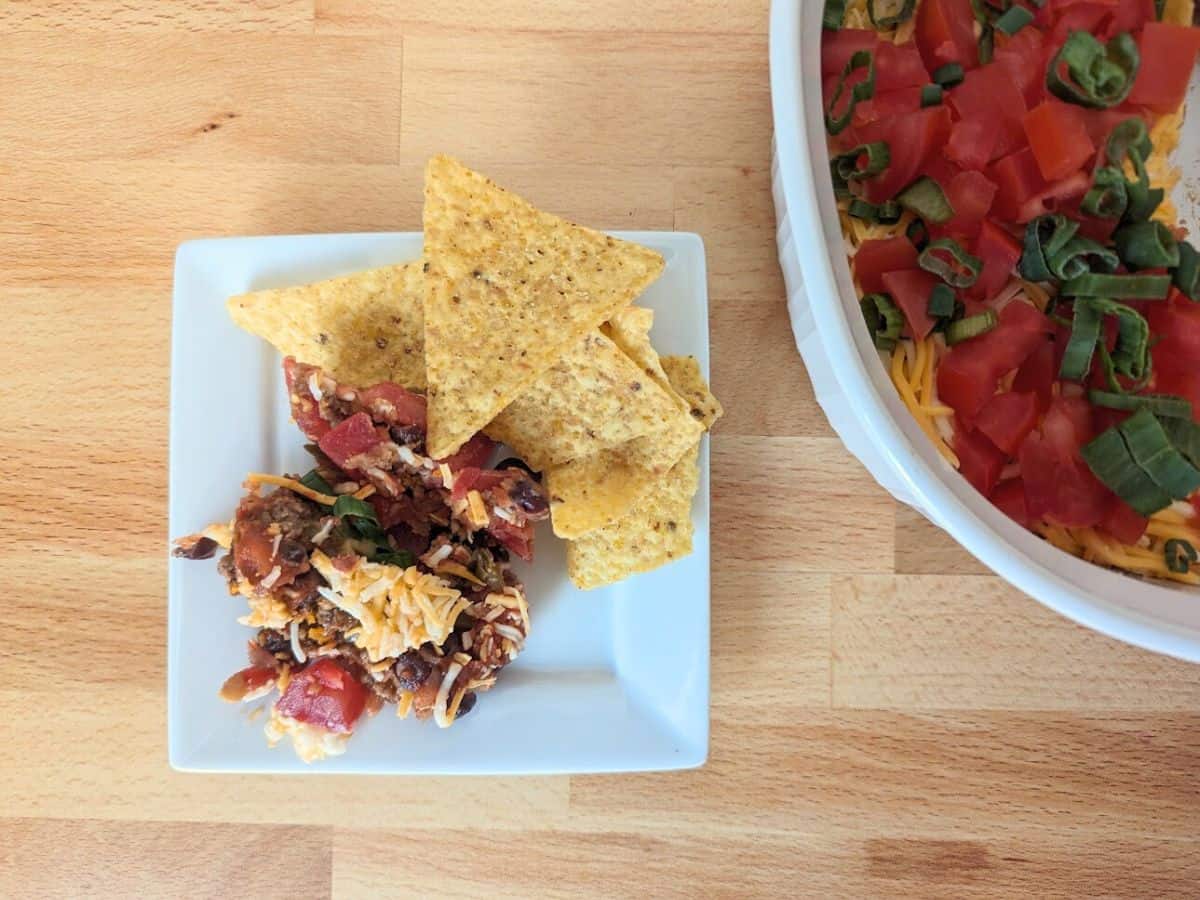 A plate with tortilla chips, black bean salsa, and shredded cheese on a wooden table next to a larger dish of the same dip garnished with diced tomatoes and green onions.