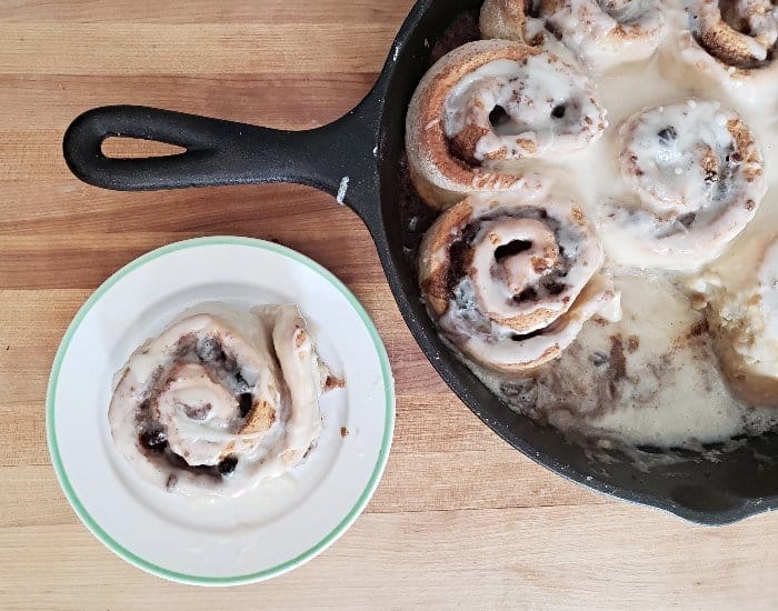 Image shows an overhead shot of a cast iron pan with cinnamon rolls and one sitting on a plate next to it.