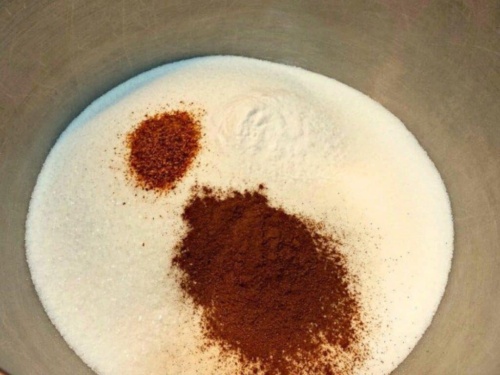 A metal bowl containing granulated sugar, ground cinnamon, ground nutmeg, baking powder, and other ingredients for Mexican sheetcake brownies, all unmixed.