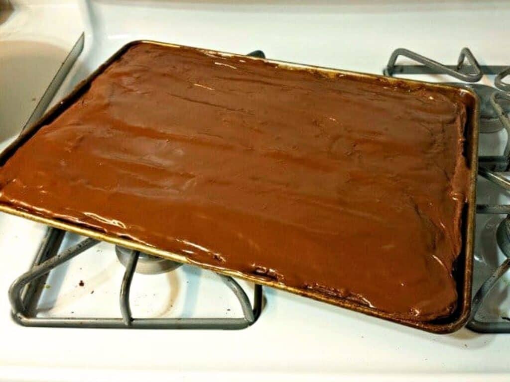 A baking sheet filled with a large, rectangular Mexican sheetcake brownie topped with smooth chocolate frosting sits on a gas stove.