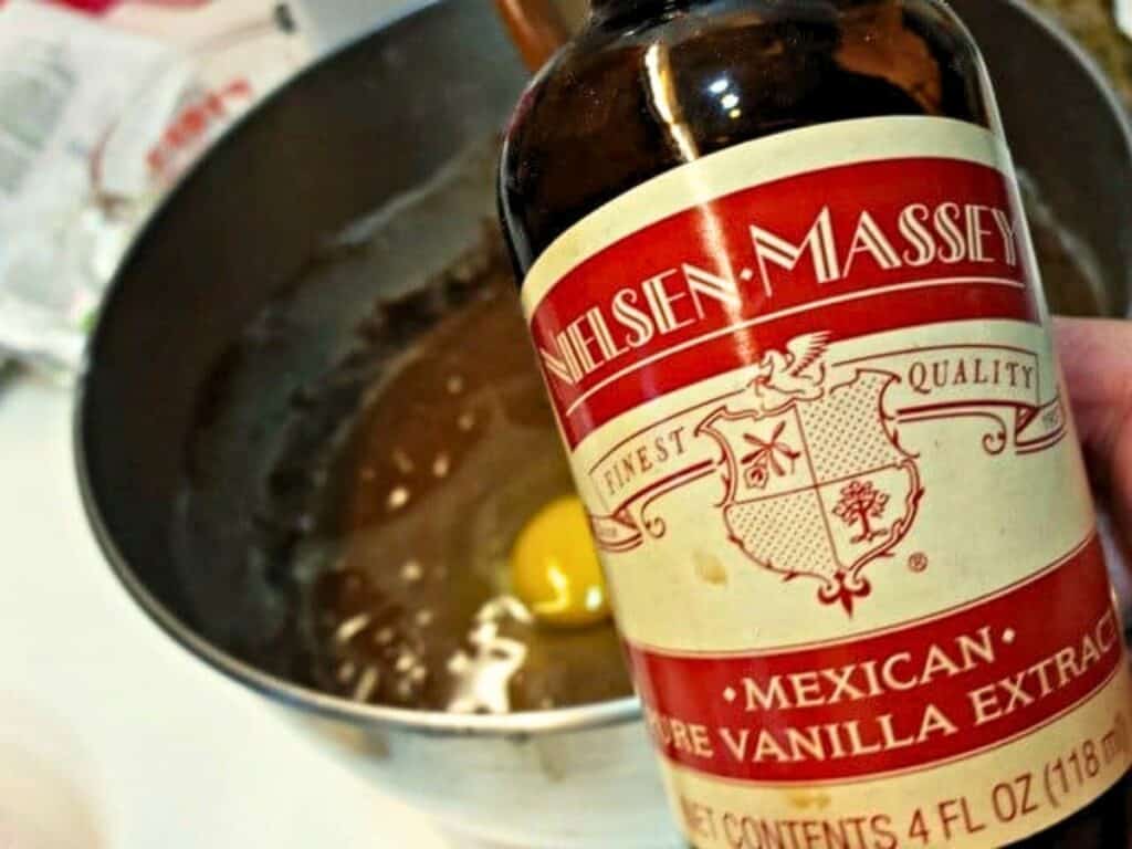 A hand holds a bottle of Nielsen-Massey Mexican vanilla extract over a mixing bowl containing batter and a whole egg, preparing the perfect base for delicious Mexican sheetcake brownies.