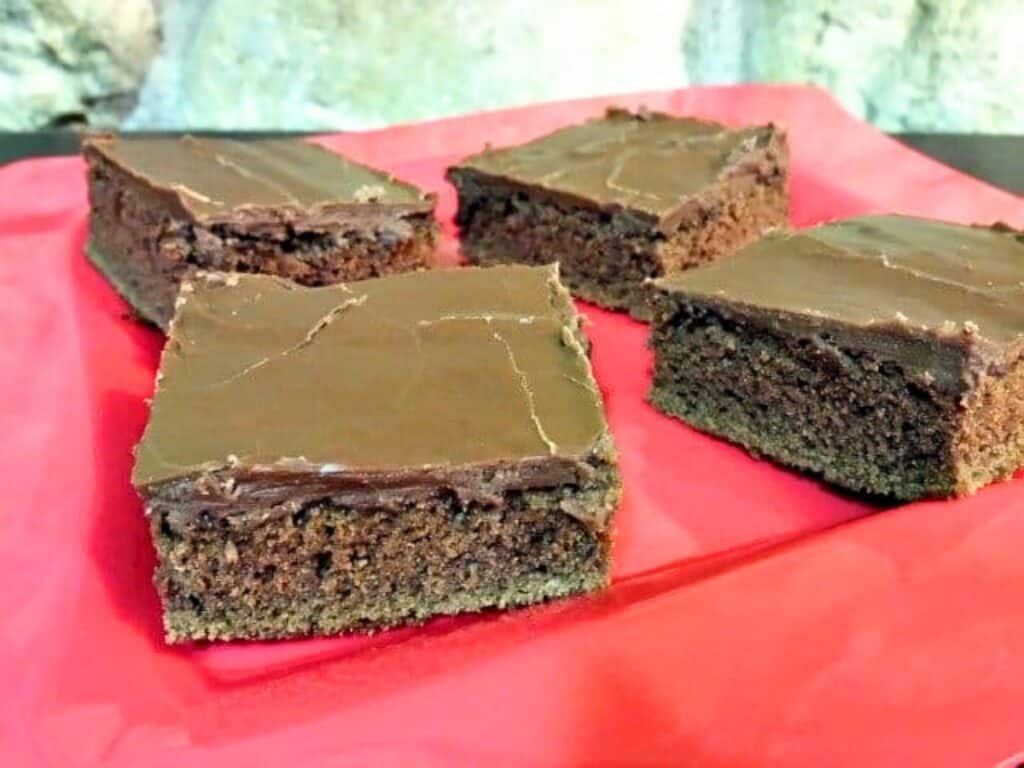 Four Mexican sheetcake brownies are arranged on a red surface, each with a smooth, glossy layer of chocolate icing on top.