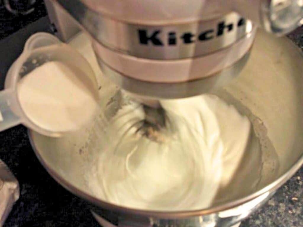 A stand mixer is whipping a white mixture in a metal bowl while sugar is being poured in from a measuring cup, the first step in creating light and airy raspberry meringues.