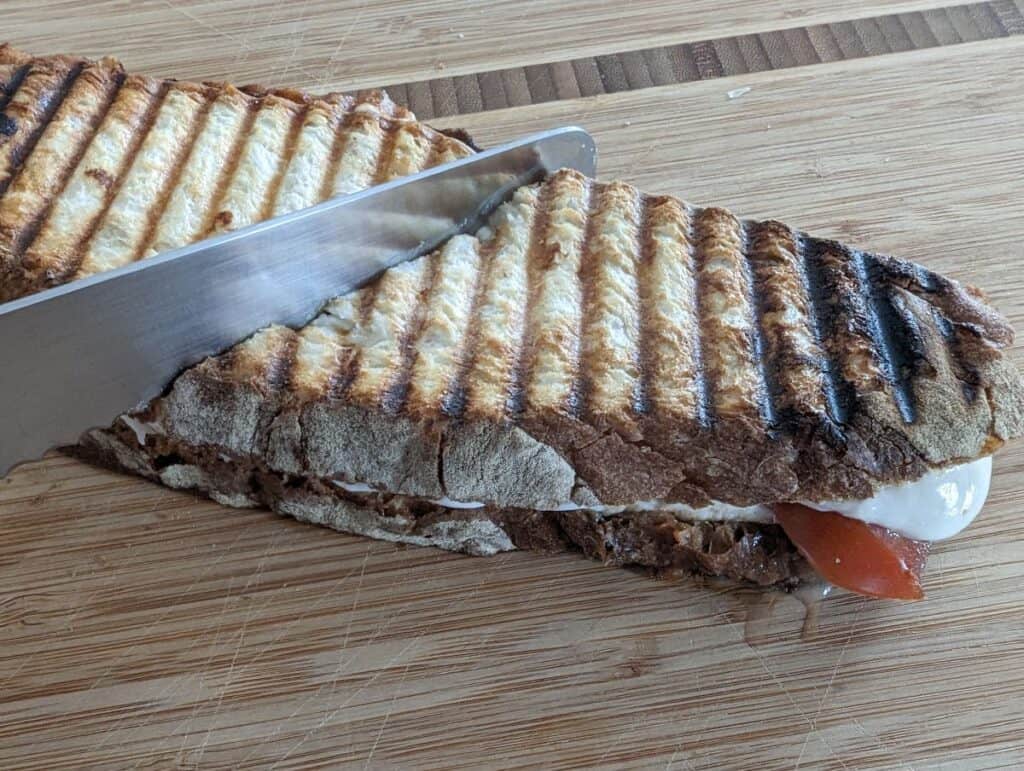 Image of a knife slicing a tomato mozzarella panini on a wooden board.