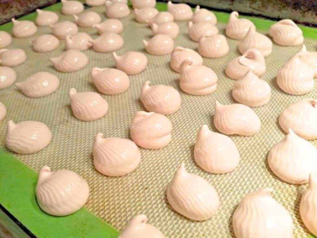 Rows of raw raspberry meringue drops, light in color, are piped onto a silicone baking mat on a tray, ready to be baked.