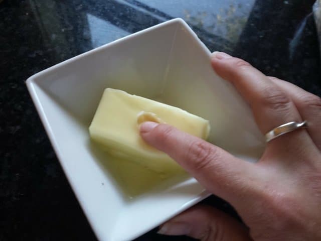Image shows a stick of butter in a white bowl with a finger testing its softness.
