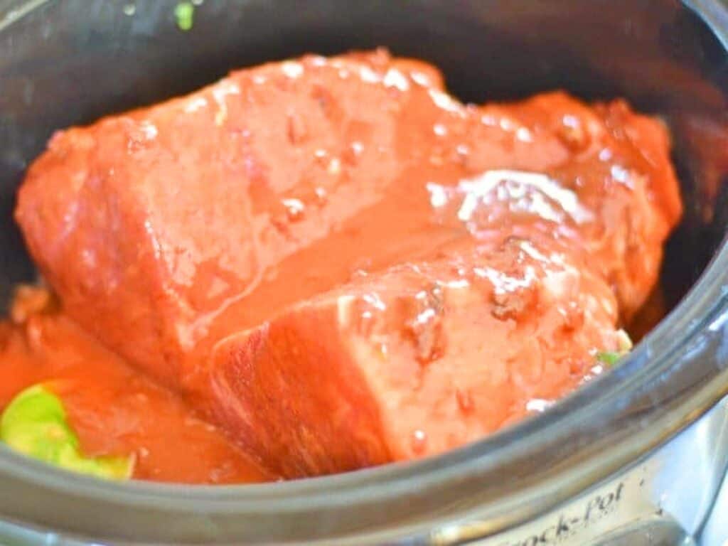 Two chunks of raw beef, perfect for crockpot Mexican beef, are covered in tomato sauce and placed inside a slow cooker, with a small piece of green vegetable visible on the side.