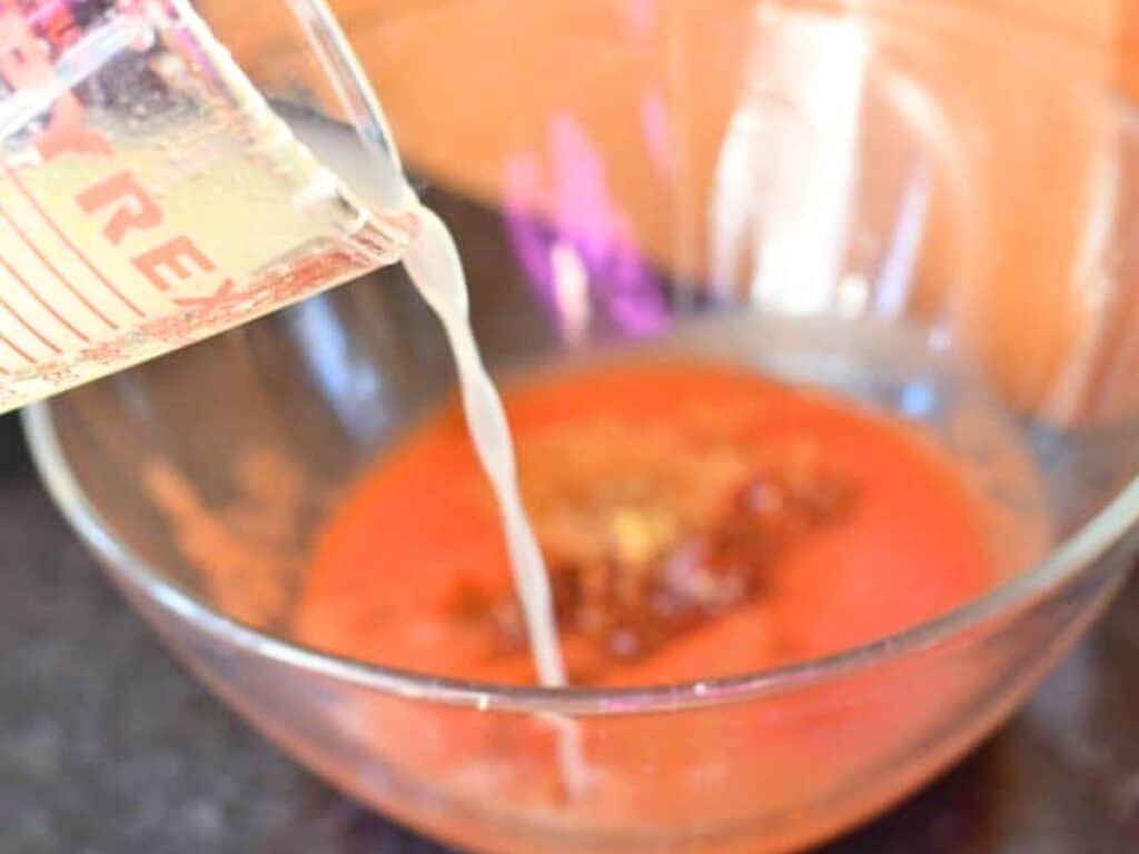 A clear measuring cup pours a liquid into a glass bowl containing a reddish mixture of spices and sauce, perfect for preparing flavorful crockpot Mexican beef.