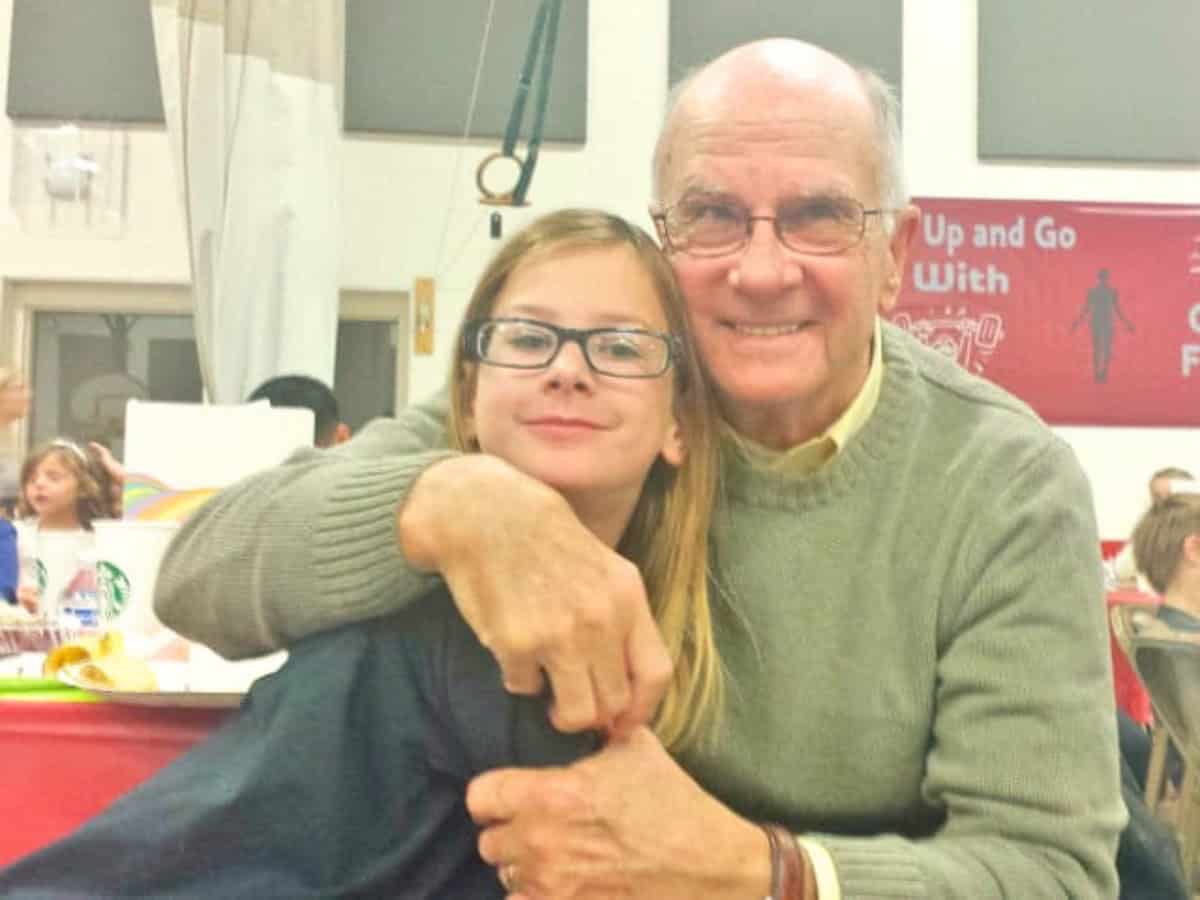 An older man wearing glasses hugs a young girl with glasses at a table in a brightly lit room, both smiling at the camera, as a delicious crockpot Mexican beef dish sits nearby.