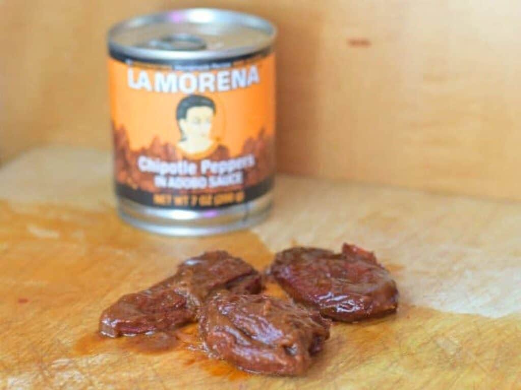 An opened can of La Morena chipotle peppers in adobo sauce is behind several chipotle peppers and sauce on a wooden surface, ready to add bold flavor to your favorite crockpot Mexican beef recipe.
