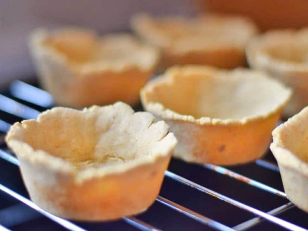 Several empty, baked mini tart shells are cooling on a metal rack, ready to be filled with savory delights like crockpot Mexican beef.