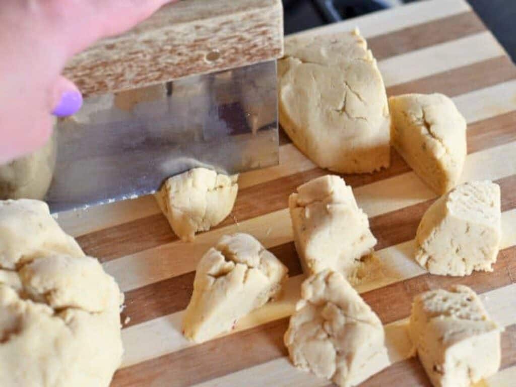 Person using a metal bench scraper to cut dough into pieces on a striped wooden cutting board, preparing the base for delicious crockpot Mexican beef empanadas.